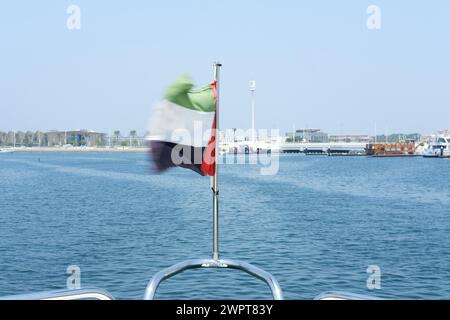 Die Flagge der Vereinigten Arabischen Emirate weht im Wind auf einer Yacht, mit der Skyline der Küste von Dubai im Hintergrund. Dubai, VAE - 15. August 2023. Stockfoto