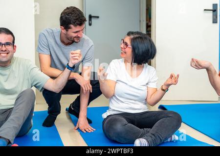 Yogalehrer spricht mit einer glücklichen erwachsenen behinderten Frau, die während des Yoga-Kurses auf einer Matte sitzt Stockfoto