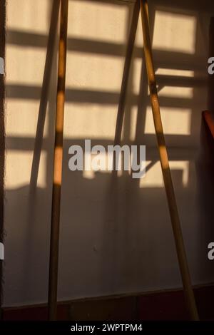 Zwei Besenstiele an einer Wand mit dem Schatten eines Fensters, das vom Licht des Sonnenuntergangs gegossen wird, in einem Haus in den östlichen Andenbergen. Stockfoto