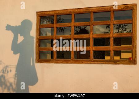 Der Schatten eines Fotografen, gegossen durch das Licht des Sonnenuntergangs an einer Wand neben einem vertäfelten Holzfenster, auf einer Farm in Zentral-Kolumbien. Stockfoto
