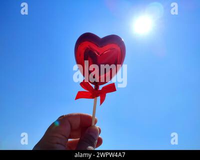 Ein Stock in Form eines volumetrischen roten Herzens in der Frauenhand vor blauem Himmel und strahlender Sonne an sonnigen Sommertagen. Concept Love, Valentinstag, verlieben, Liebste. Vertikal Stockfoto