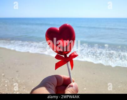 Ein Stock in Form eines volumetrischen roten Herzens in der Frauenhand vor dem Hintergrund des blauen Meeres, des Sandstrandes und des blauen Himmels an sonnigen Sommertagen. Concept Love, Valentinstag, Verlieben. Vertikal Stockfoto