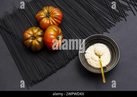 Drei saftige, reife Raf-Tomaten auf einem Hügel schwarzer Nudeln neben einer Schüssel mit geriebenem Käse und einem goldenen Löffel Stockfoto