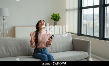 Überglückte Frau Mädchen Frau auf Sofa benutzen Handy aufgeregt Gewinner Kunde erhalten E-Commerce Shopping Angebot gewinnen Bid Jump lustig tanzen auf Stockfoto