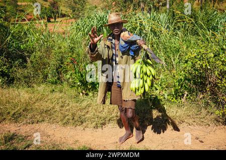 Sendrisoa, Madagaskar - 27. April 2019: Unbekannter madagassischer Mann in Strohhut und Mantel, barfuß auf staubiger Straße, Bananen auf hölzernem Pfosten Stockfoto