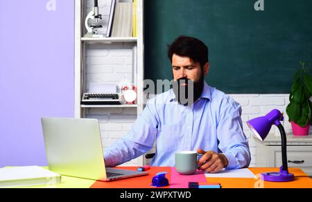 Männlicher Lehrer oder Schüler, der einen Laptop benutzt, der am Schreibtisch sitzt und online studiert. Schullehrer. Virtuelle Bildungskurse. Bärtiger Lehrer, der sich auf den Test vorbereitet oder Stockfoto