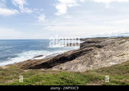 Felsige Küste mit Grün, mit Blick auf ein riesiges, ruhiges Meer unter einem teilweise bewölkten Himmel. Stockfoto