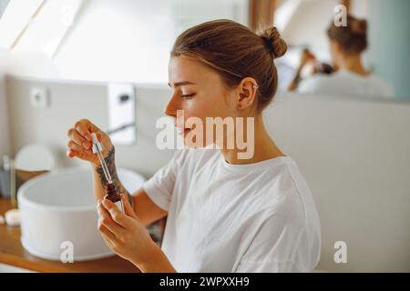 Schöne junge Frau, die im Badezimmer Kosmetikserum auf ihr Gesicht aufträgt. Gesichtspflege Stockfoto