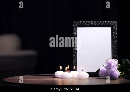 Trauerfotorahmen mit schwarzem Band, violetten Rosen und brennenden Kerzen auf Holztisch drinnen. Leerzeichen für Text Stockfoto