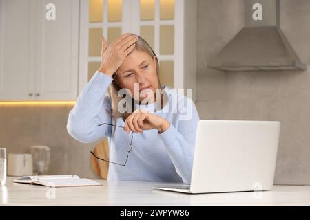 Wechseljahre. Frau mit Brille, die an Kopfschmerzen am Tisch in der Küche leidet Stockfoto