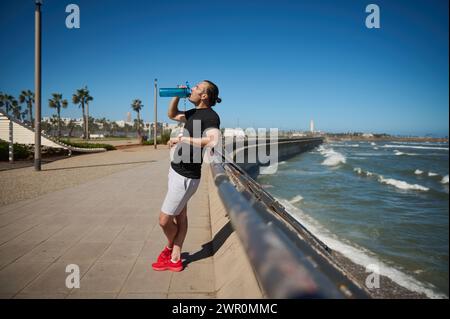 Lifestyle-Porträt in voller Länge junger Sportler, Sportler in Aktivkleidung, Trinkwasser nach dem Training im Freien. Männlicher Athlet im Ruhezustand nach Sport tr Stockfoto