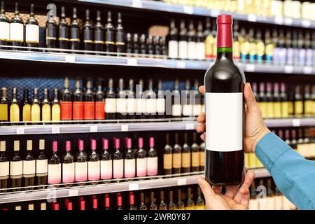 Weinflasche mit leerem Etikett in den Händen des Mannes vor Weinflaschen in Regalen im Spirituosengeschäft. Stockfoto