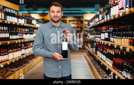 Fröhlicher Kundenmann, der eine Flasche Wein in der Hand hält und im Spirituosenladen lächelnd. Stockfoto