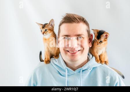 Lächelnder lustiger Mann in blauem Hoodie mit zwei Abessinierkatzen, kleinen neugeborenen Kätzchen, die auf Schultern sitzen. Weißer Hintergrund. Glückliche Haustiere, flauschige Haustiere Stockfoto