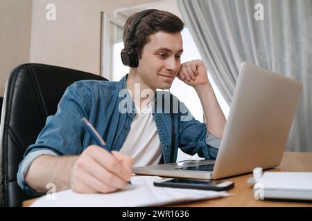 Glücklicher Schüler, der Kopfhörer mit Laptop für Fernunterricht trägt, Videokonferenzen genießt, Webinar ansieht, Audiokurse anhört, Notizen schreibt, mit dem Lehrer lernt, Lektion haben Stockfoto
