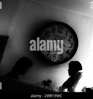 Schwarzweißbild in einem Café mit zwei Personen, die sich bei Kaffee unterhalten, mit einer großen antiken Uhr an der Wand, Großbritannien Stockfoto