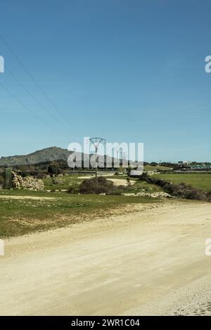 Ein ländlicher Feldweg, flankiert von Grasfeldern und einer Stromleitung auf Hochmasten Stockfoto