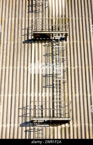 Ein Blick auf eine Reihe von gestaffelten geschützten Leitern, die das Ende eines großen Lagerhausgebäudes auf einem Industriegebiet hinaufsteigen. Stockfoto