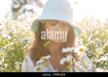 Mädchen, das auf einer Wiese sitzt, die mit wilden Blumen bedeckt ist. Hintergrund des Blühfeldes der Gänseblümchen. Sonniger Sommertag, Lichtstrahlen. Sommerferienkonzept Stockfoto