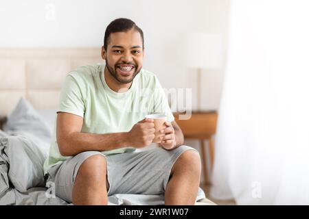 Lächelnder, gutaussehender schwarzer Mann, der morgens Kaffee genießt, Schlafzimmer-Innenausstattung Stockfoto