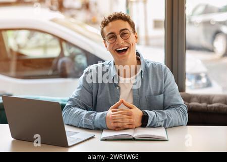 Lachender junger Mann mit lockigen Haaren und Brille, der mit Händen vor seinem offenen Notizbuch sitzt Stockfoto