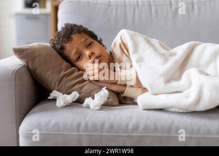 Der kranke kleine schwarze Junge ruht sich zu Hause unter der Decke auf der Couch aus Stockfoto