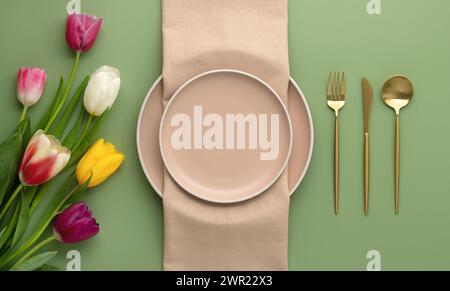 Festliche Atmosphäre mit beiger Serviette und Frühlingsblumen. Leere Teller und goldenes Besteck auf grünem Hintergrund. Draufsicht. Esstisch im luxuriösen Restaurant Stockfoto