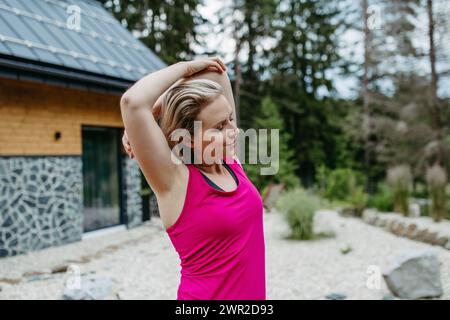 Junge Frau, die sich vor dem Training ausdehnt, im Wald läuft, sich auf einer von der Natur umgebenen Terrasse des Berghauses vorbereitet. Stockfoto