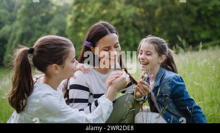 Schöne Mutter mit Töchtern, Blumen pflücken, Haare einziehen, im Gras auf der Wiese sitzen. Muttertag und mütterliche Liebe. Stockfoto