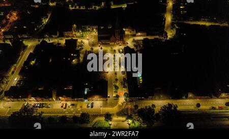 Dieses nächtliche Luftbild fängt die Umgebungsbeleuchtung eines Stadtplatzes ein, wo das warme Leuchten der Straßenlaternen ein Netz von Sichtbarkeit vor dem dunklen Hintergrund der Nacht erzeugt. Der zentrale Platz, der möglicherweise durch eine Kirche oder ein bedeutendes Gebäude verankert ist, wird mit seiner Umgebung in künstlichem Licht zum Mittelpunkt. Die Scheinwerfer und Rücklichter des Fahrzeugs ziehen sich durch die Straßen und folgen den Pfaden der späten Abendaktivitäten. Die dunklen Silhouetten von Bäumen und die umgebende Architektur bilden den Rahmen der Szene und geben ein Gefühl des ruhigen Lebens nach der Dämmerung in einer urbanen Umgebung. Dies Stockfoto