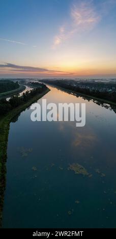 Dieses Bild zeigt die ruhige Pracht des Sonnenaufgangs, als es über einen ruhigen Wasserweg bricht. Der Himmel, der in Pastelltönen in Rosa, Blau und Gold gemalt ist, spiegelt sich perfekt von der Wasseroberfläche wider und schafft einen symmetrischen Farbteppich mit Licht und Farbe. Wolken verstreut über den Himmel und verleihen der ruhigen Szene Textur. Das Vorhandensein von Grün, das auf der Wasseroberfläche schwimmt, bringt einen Hauch von Leben und Unvollkommenheiten der Natur in die ansonsten ruhigen Gewässer und deutet auf das Ökosystem im Inneren hin. Die Böschungen umrahmen den Wasserweg und führen den Blick des Betrachters zum Horizont und zum Versprechen des Stockfoto