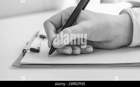 Schwarzweißfoto. Konzentriere dich auf eine weibliche Hand, die einen Stift hält, um etwas auf ein leeres Blatt Papier zu schreiben. Stockfoto