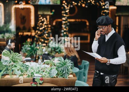 Ein modisch gekleideter Afroamerikaner spricht am Telefon, während er Dokumente in einem Café hält, das mit festlichen Weihnachtslichtern geschmückt ist. Stockfoto