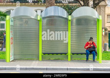 Frau in rot gekleidet mit ihrem Handy an einer grünen Bushaltestelle. Portugalete-Baskenland-Spanien. 3-3-2024 Stockfoto