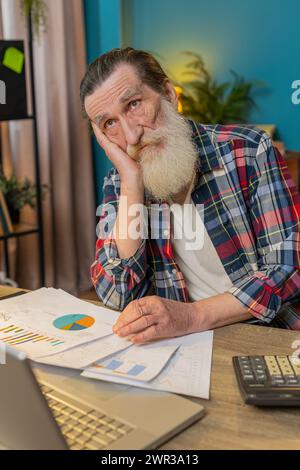 Müde erschöpfter Senior man Freelancer, der Papierkramme analysiert, Zahlungen abrechnet und monatliche Ausgaben für Bankrechnungen am Home Office Desk berechnet. Verärgerter reifer Opa, der am Tisch arbeitet. Stockfoto