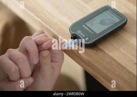 Blutzuckermessgerät mit Teststreifen, die auf einem Holztisch liegen, Kinderhand hält einen Finger und führt ihn zum Messgerät, Blutzuckermessung Stockfoto