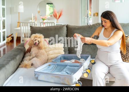 Schwangere Frau, die einen Koffer vorbereitet, um ins Krankenhaus zu gehen, auf einem Sofa neben einem Hund sitzend Stockfoto