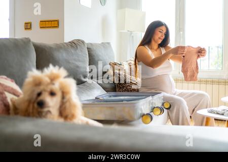 Glückliche schwangere Frau, die sich bereit macht, ins Krankenhaus zu gehen, Kleidung und Koffer vorzubereiten, die auf dem Sofa neben einem Hund sitzen Stockfoto