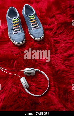 Blaue Sneakers mit bunten Schnürsenkeln und weißen Kopfhörern mit Kabelanschluss auf roter Haaroberfläche Stockfoto