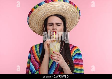 Junge Frau mit mexikanischem Sombrero-Hut und Poncho, der Cocktail auf rosa Hintergrund trinkt Stockfoto