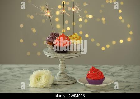 Verschiedene bunte Cupcakes mit Glitzern auf weißem Marmortisch vor verschwommenem Licht Stockfoto