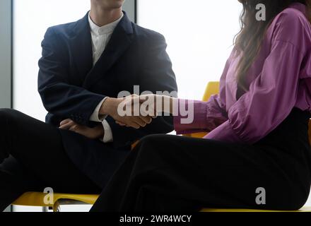 Zwei Leute schütteln sich die Hände in einem Geschäftsumfeld. Der Mann trägt Anzug und Krawatte, während die Frau ein lila Hemd trägt Stockfoto