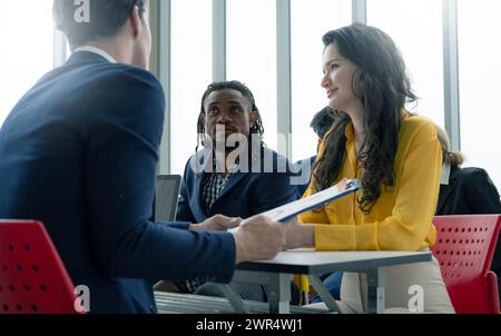 Eine Gruppe von Personen sitzt um einen Tisch, von denen einer eine eine Zwischenablage hält. Die Atmosphäre scheint professionell und ernst zu sein, so wie die Gruppe Stockfoto
