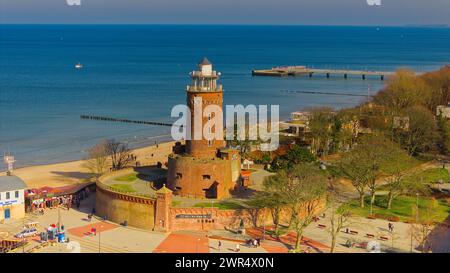 Februar in Kołobrzeg, Polen: Eine ruhige Szene mit einem Leuchtturm aus rotem Backstein, Menschen, die einen sonnigen Tag am Sandstrand genießen, und einem ruhigen Meer mit Stockfoto