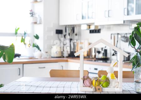 Schlüssel und winziges Haus mit gemütlichem Haus mit Osterdekor mit Kaninchen und Eiern auf dem Tisch der Küche. Bau, Entwurf, Projekt, Umzug in ein neues Haus, Hypothek, Stockfoto