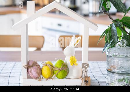 Schlüssel und winziges Haus mit gemütlichem Haus mit Osterdekor mit Kaninchen und Eiern auf dem Tisch der Küche. Bau, Entwurf, Projekt, Umzug in ein neues Haus, Hypothek, Stockfoto