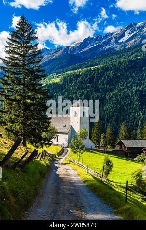 Eingebettet in hoch aufragende grüne Berge, ragt eine kleine weiße Kirche mit einem markanten Glockenturm in einem ruhigen Bergdorf hervor. Umgeben von Stift Stockfoto