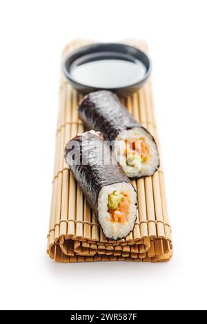 Eine Bambusmatte mit zwei Sushi-Brötchen, die ordentlich neben einer Schüssel Sojasauce platziert werden. Die Sushi-Brötchen sind mit Reis, Algen und verschiedenen Zutaten gefüllt. Stockfoto