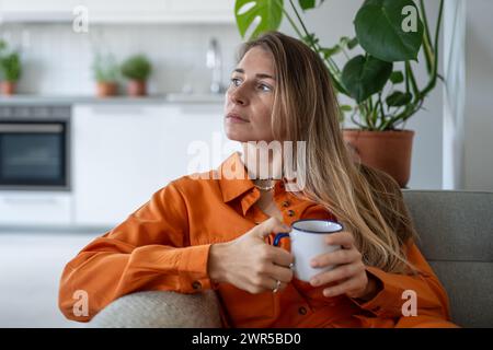Träumende Frau mittleren Alters, die mit einer Tasse Tee auf dem Sofa sitzt und sich zur Seite schaut, denkt nachdenklich, visualisiert Stockfoto
