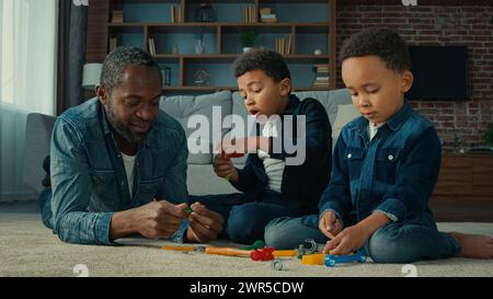 Afroamerikaner ethnischer Mann Vater mit zwei kleinen Vorschulsöhnen Kinder spielen Spiel auf dem Boden zu Hause, Vater mit Kindern, die mit Werkzeugen spielen Spielzeug Stockfoto